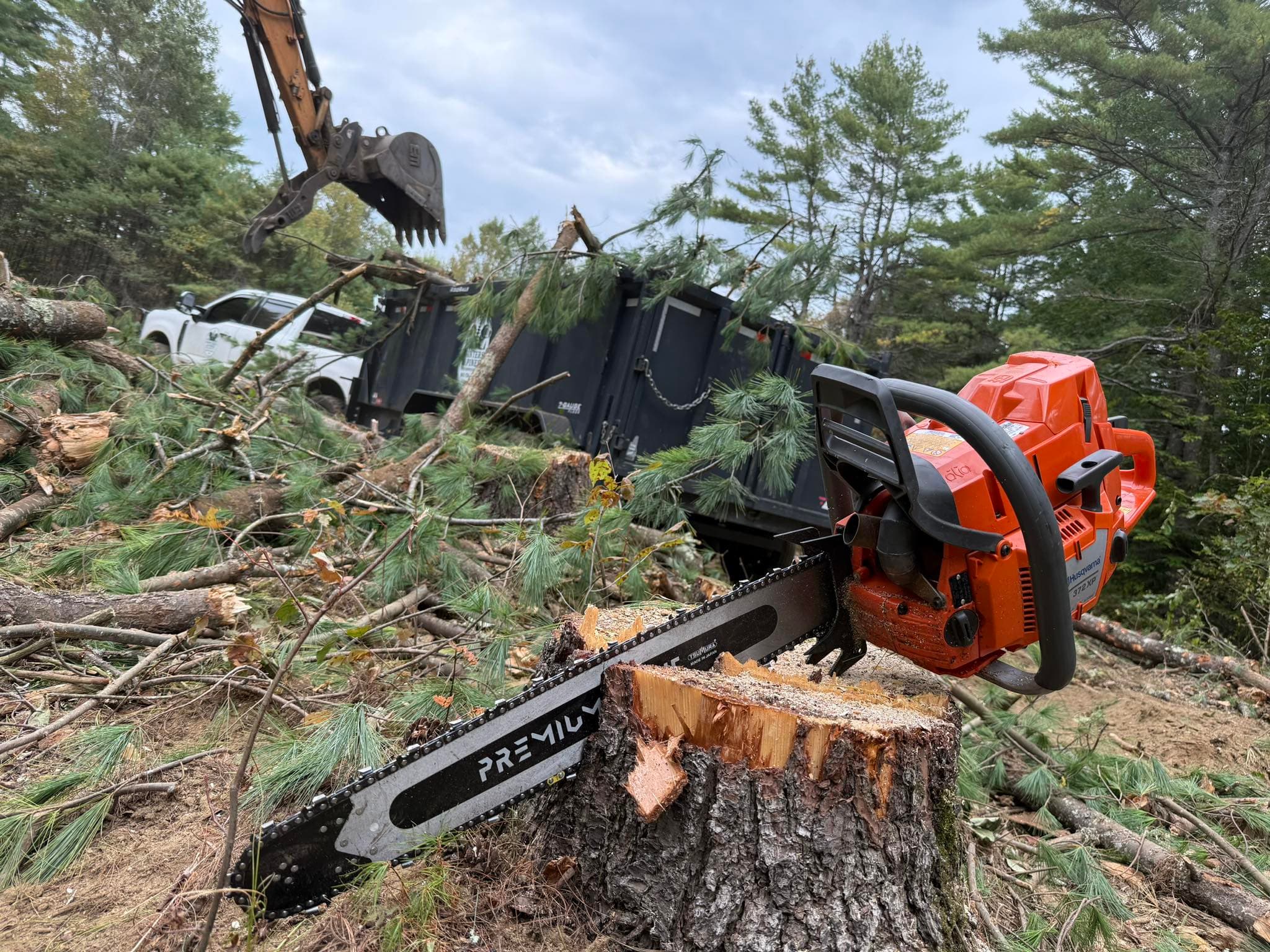 Orange chainsaw on a tree stump with an excavator loading branches into a trailer.